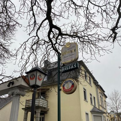 Ein Restaurant-Schild mit der Aufschrift "Hellas" steht an einem Gebäude. Die Äste eines kahlen Baumes sind im Vordergrund sichtbar.