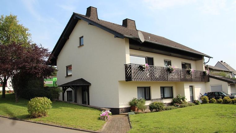 A large, two-story house with a balcony and flowers. The garden is well-maintained and there are some trees and shrubs.
