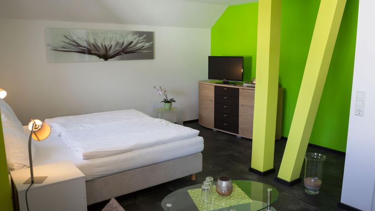 Ein modernes Zimmer mit einem Doppelbett und klaren Linien. Die Wände sind grün gestrichen, und es gibt einen Fernseher sowie eine Kommode.