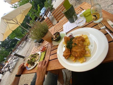 Ein Tisch im Freien mit zwei Tellern Essen. Auf einem Teller ist ein paniertes Schnitzel mit Kartoffeln, und der andere Teller enthält einen bunten Salat.