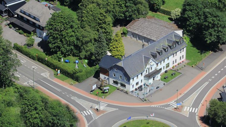 Eine Vogelperspektive auf einen Kreisverkehr mit einem weißen Gebäude und viel Grün umgeben. Man sieht auch einige Autos und Wege im Bild.
