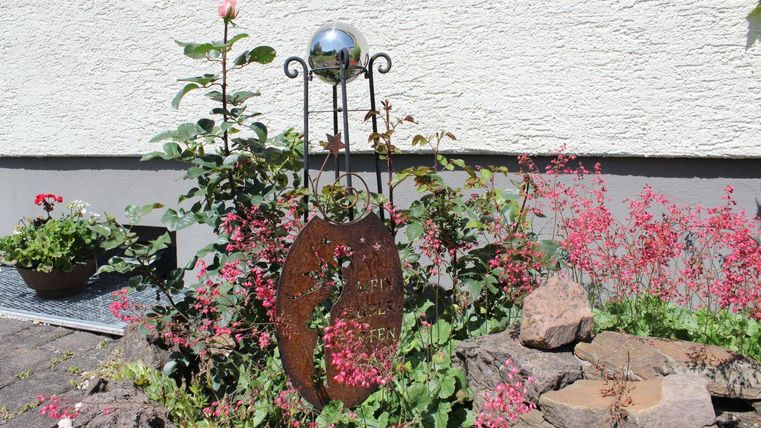 A decorative garden spot with a metal panel and many colorful flowers. In the background, a bright wall is visible.