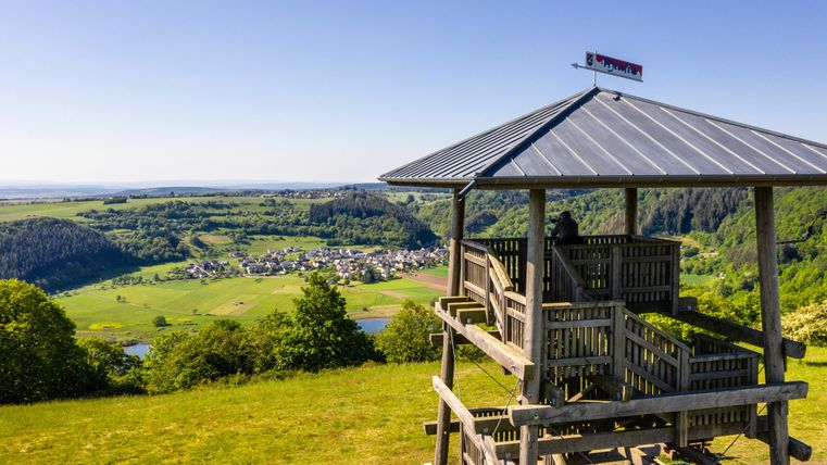 Ein Aussichtsturm auf einem Hügel mit Blick auf eine grüne Landschaft und ein kleines Dorf im Tal. Der Himmel ist klar und blau, ideal für einen schönen Ausblick.