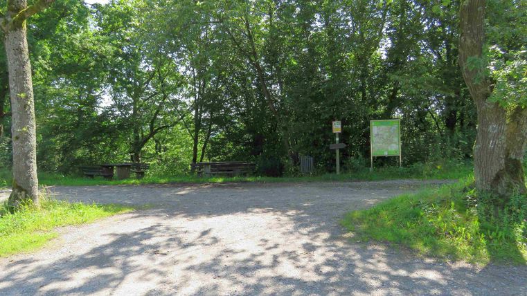 Eine ruhige Weggabelung im Grünen, umgeben von Bäumen. Ein Schild deutet auf einen möglichen Wanderweg hin.