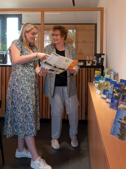 Zwei Frauen stehen in einem modernen Raum und schauen sich gemeinsam Broschüren an. Im Hintergrund ist eine Auslage mit verschiedenen Informationsmaterialien zu sehen.