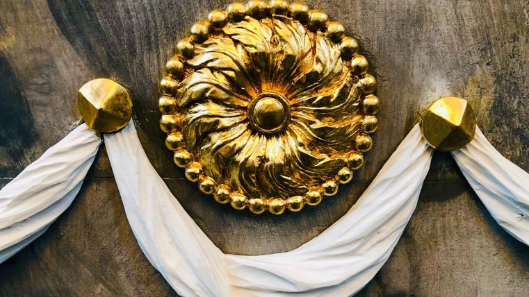 Eine goldene Dekoration mit einem floralen Muster ist in der Mitte zu sehen. An den Seiten hängen weiße Drapierungen.