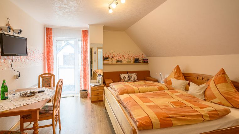 Ein gemütliches Zimmer mit zwei Betten und orangefarbener Bettwäsche. Im Hintergrund befindet sich ein Tisch mit Stühlen und ein Fernseher an der Wand.