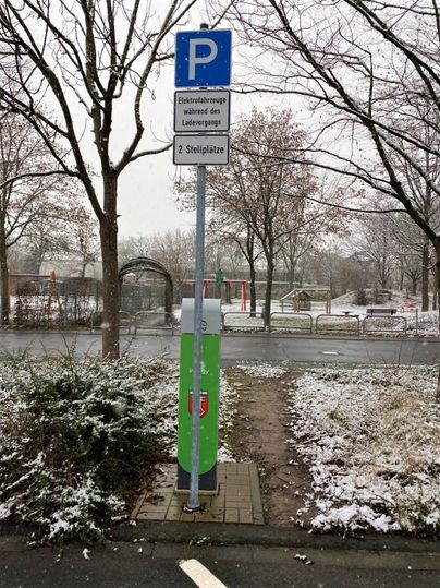 A parking space for electric vehicles with a charging station sign. In the background, trees and a wintry landscape can be seen.