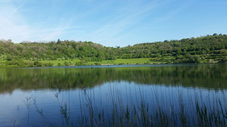 Ein ruhiger See, umgeben von grünen Hügeln und Bäumen. Der Himmel ist klar und blau, und das Wasser spiegelt die Landschaft wider.