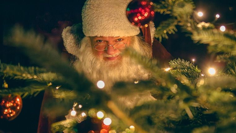 Ein freundlicher Weihnachtsmann blickt zwischen Tannenzweigen hervor. Die Szene ist festlich beleuchtet mit bunten Weihnachtskugeln.