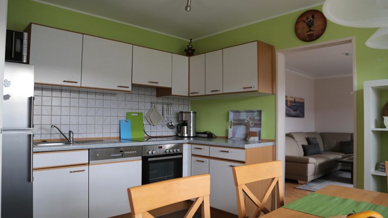 Eine moderne Küche mit grünen Wänden und weißen Schränken. Im Vordergrund steht ein Esstisch mit Holzstühlen.