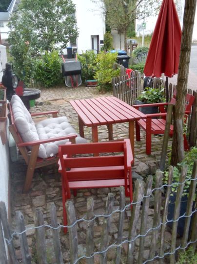 Een gezellige zithoek buiten met rode meubels en een parasol. Omgeven door planten en een klein hek.