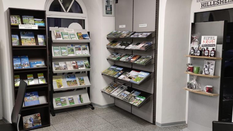 Innenansicht eines Tourismusbüros in Hillesheim mit Regalen voller Broschüren und Souvenirs. Ein Schild weist auf den Eifel Krimi Wanderweg hin.