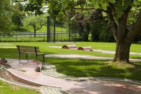 A beautiful mini-golf course in a park with green grass and shady trees. In the background, a bench and a sports field can be seen.