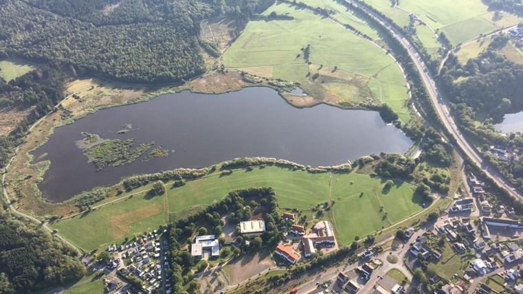Een uitgestrekt landschap met een rustige meer, omringd door groene weilanden en bomen. Op de voorgrond zijn enkele gebouwen en wegen zichtbaar.