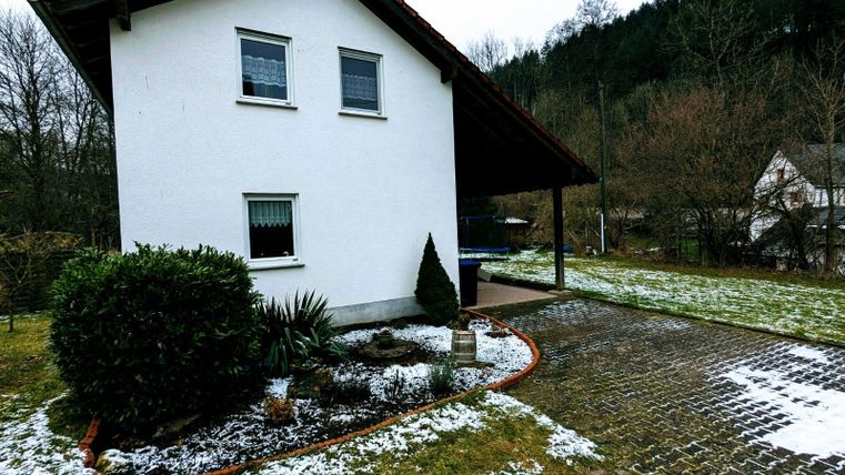 A white house with a red roof and a small front yard. The ground is partially covered with snow and there are trees in the background.