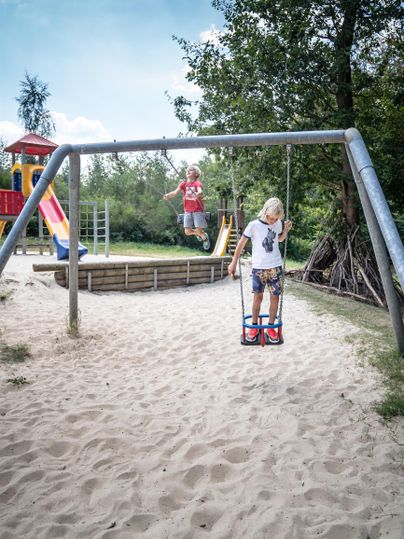 Ein Kind schwingt fröhlich auf einer Schaukel in einem Sandspielplatz. Im Hintergrund sind Rutschen und Spielgeräte zu sehen.