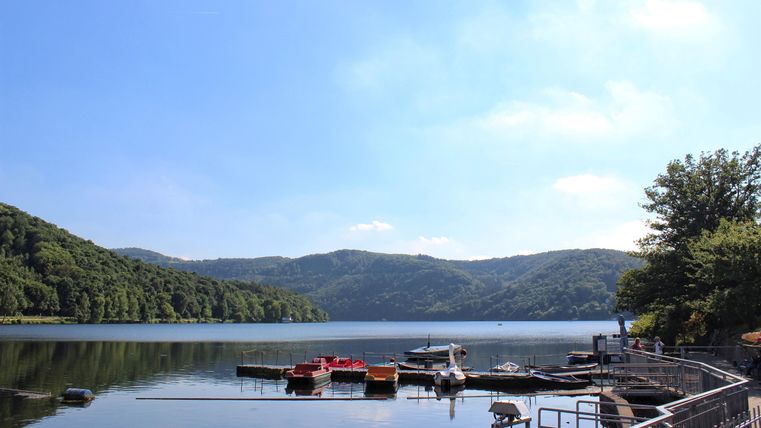 Ein ruhiger See umgeben von sanften Hügeln und grünem Wald. Am Ufer liegen Bootsanleger und Boote, die auf das Wasser warten.