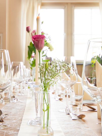 A festively laid table with glasses and flowers in a vase. In the background, candlelight and a bright room are visible.