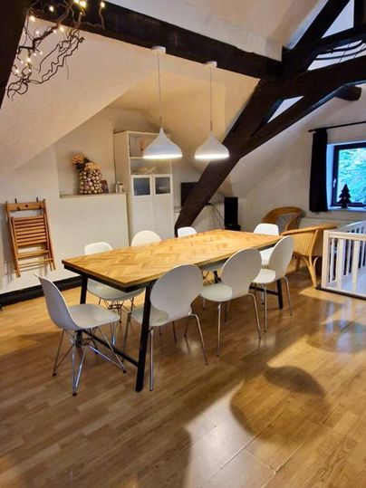 A cozy dining area with a long wooden table and white chairs. The room has high ceilings and visible beams, as well as warm light from hanging lamps.