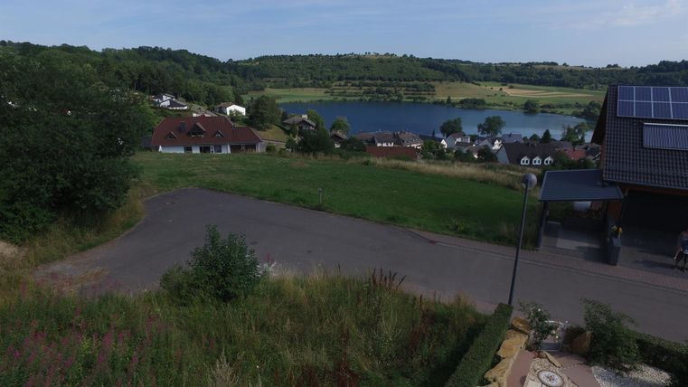 Eine malerische Landschaft mit einem ruhigen See und sanften Hügeln. Im Vordergrund ist eine Wiese und eine Straße, umgeben von Häusern.