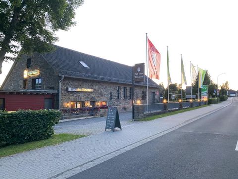 A cozy restaurant in a stone building with various flags in front of the entrance. The surroundings display a quiet street and green trees.