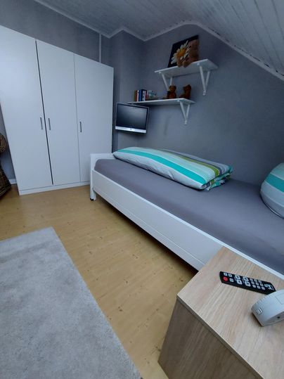 A simple room with a single bed, a TV, and a wardrobe. The floor is wooden and a small table stands next to the bed.