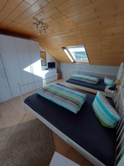 A cozy bedroom with two single beds and colorful pillows. Large windows provide plenty of daylight and a view outside.