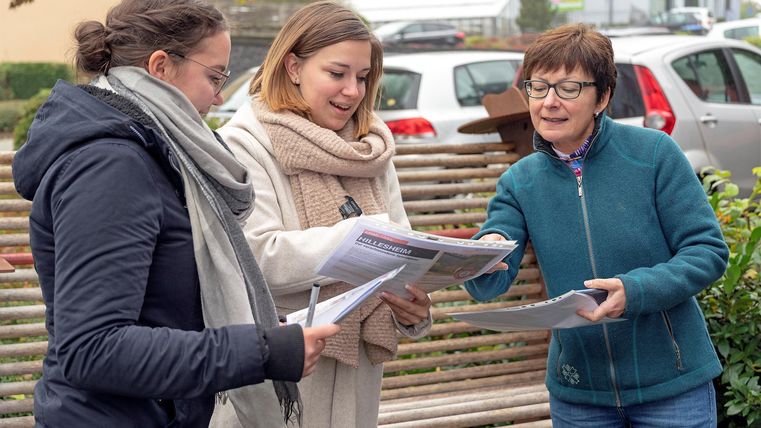 Three women are standing outside and looking at a document together.