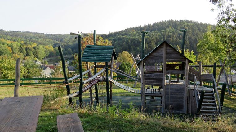 Een speeltuin met houten constructies en een glijbaan in een natuurlijke omgeving. Op de achtergrond zijn bomen en zachte heuvels te zien.