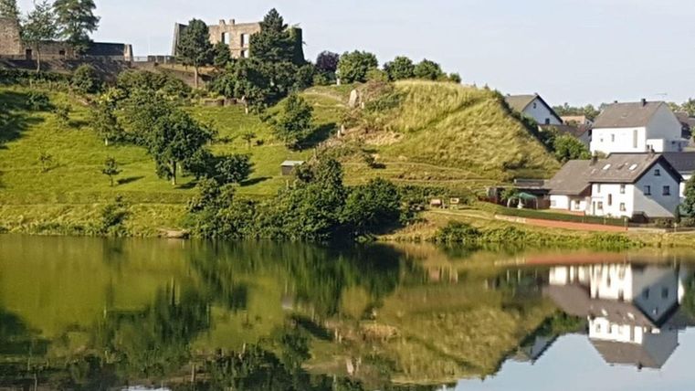 Een schilderachtig landschap met een rustige meer en groene heuvels. Op de achtergrond zijn historische gebouwen en kleinere huizen te zien.
