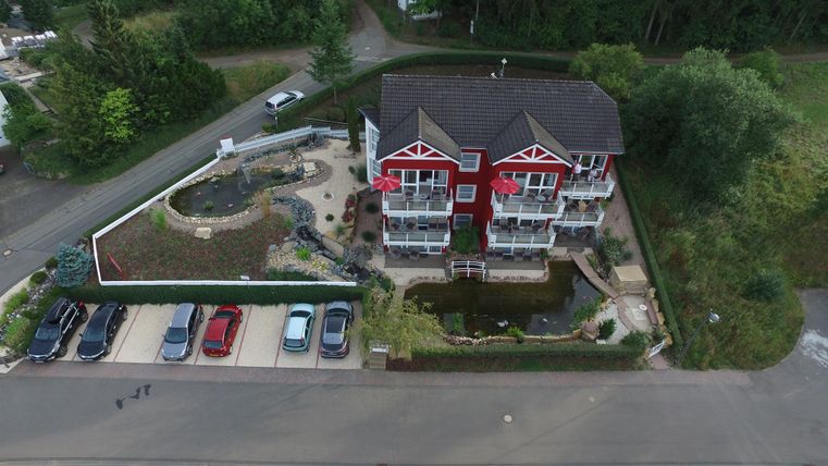 Ein modernes Gebäude mit roten Akzenten und mehreren Etagen. Darum herum befinden sich parkende Autos und ein gepflegter Garten mit einem kleinen Teich.