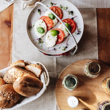 Ein gedeckter Tisch mit frischem Brot und einer Schüssel Tomaten und Mozzarella. Darauf befinden sich frische Basilikumblätter und Gewürze in kleinen Gläsern.