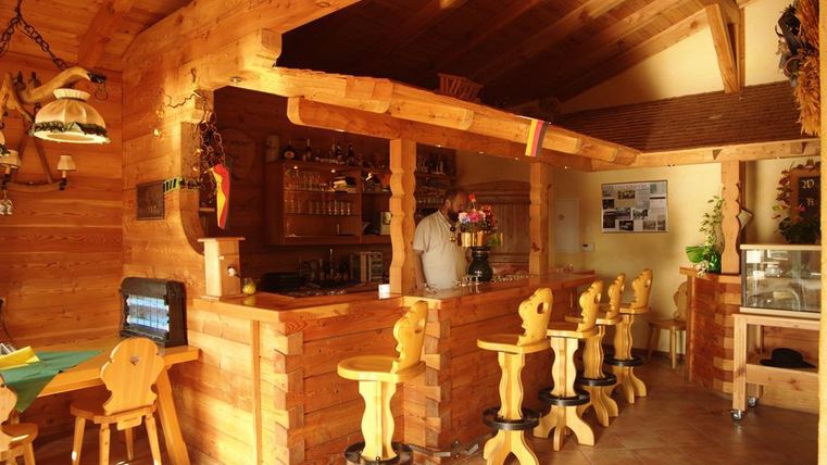 Ein gemütliches Holzinterieur mit einer Theke und Hockern. Im Hintergrund sind Getränke und Dekorationen sichtbar.