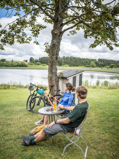Drie personen zitten onder een boom aan de oever van een water. Op de achtergrond staan fietsen en het landschap is groen en bosrijk.
