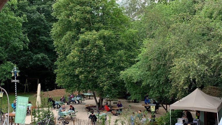 Een gezellige buitenruimte met veel bomen en tafels. Mensen zitten te genieten van de natuur.