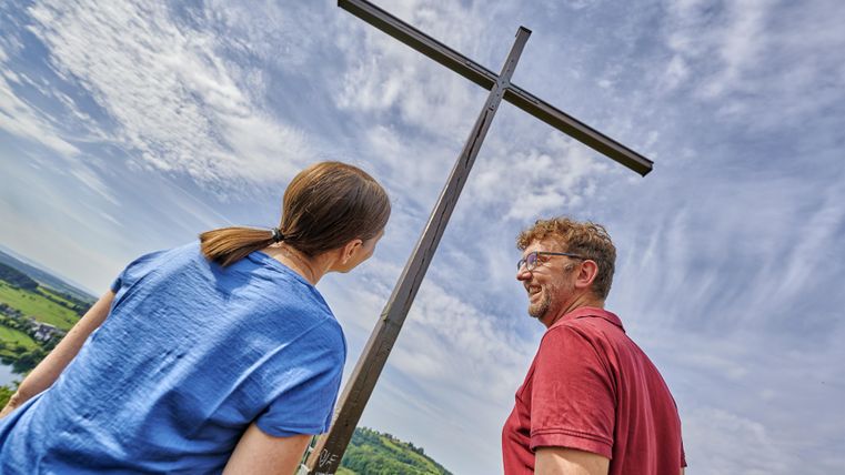 Zwei Personen schauen zu einem großen Holzkreuz hinauf. Der Himmel ist überall mit schönen Wolken geschmückt.