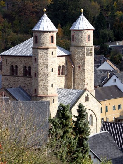 Een historische kerk met twee torens en een gewelfd dak. Omgeven door een schilderachtig landschap en woongebouwen.