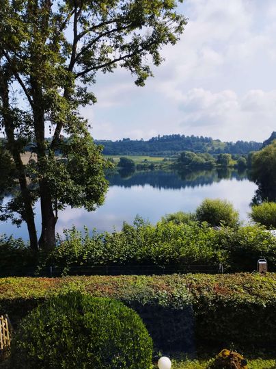 Eine ruhige Landschaft mit einem klaren See und grünen Bäumen. Im Hintergrund sind sanfte Hügel unter einem blauen Himmel zu sehen.