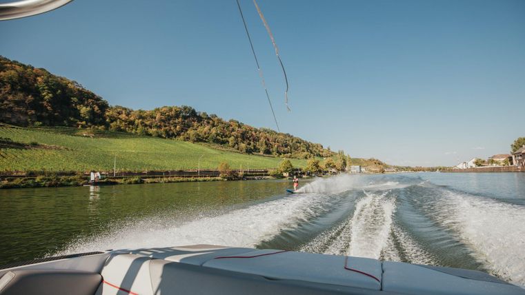 A boat is sailing on a calm body of water, surrounded by gentle hills and green meadows. The sky is clear and blue.