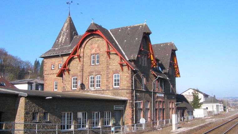 View of the back of the brick railway station building from the platforms. You can see the flat annex and the three-story main building with the inscription Jünkerath. In the foreground, the railway tracks are visible.