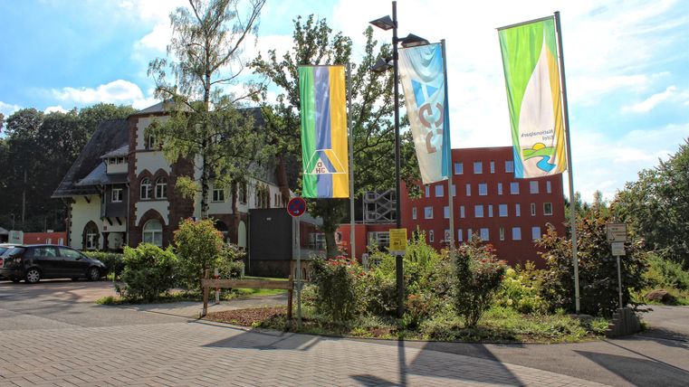 Ein Bereich mit mehreren Fahnen und zwei Gebäuden im Hintergrund. Der Himmel ist klar und sonnig.