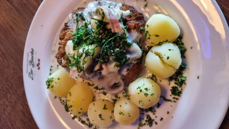 Een heerlijk gerecht met een schnitzelachtig hoofdgedeelte, bedekt met een witte saus en verse kruiden. Daarbij worden gekookte aardappelen geserveerd, die keurig zijn opgediend.