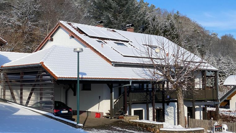 Ein modernes Haus mit einem schneebedeckten Dach. Im Vordergrund liegt frischer Schnee und es ist ein klarer blauer Himmel zu sehen.