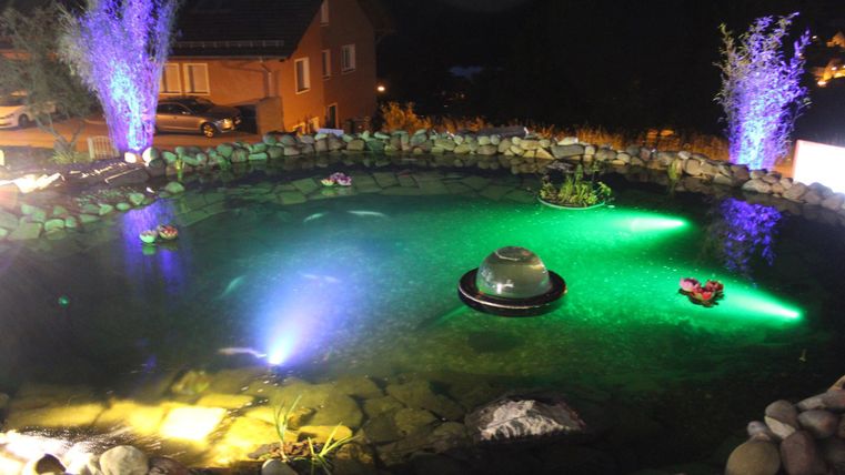 Ein schöner Teich bei Nacht, beleuchtet mit bunten Lichtern. Umgeben von Steinen und ein paar Pflanzen.