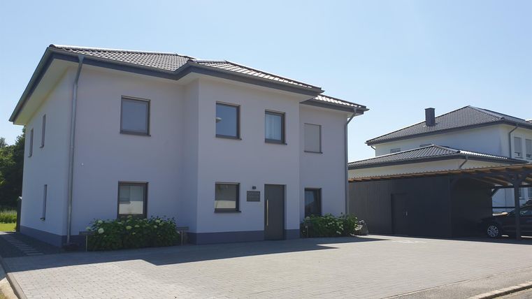 Ein modernes, zweigeschossiges Haus in heller Farbe. Es hat eine asphaltierte Einfahrt und einen klaren, blauen Himmel im Hintergrund.