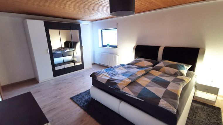 A modern bedroom with a large bed and a mirrored wardrobe. The room has wooden flooring and soft lighting.