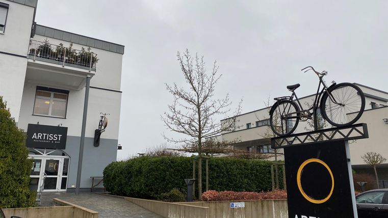 Ein modernes Gebäude mit einem Schild, das "ARTISST" zeigt. Vor dem Gebäude steht ein Gestell mit einem Fahrrad darauf.
