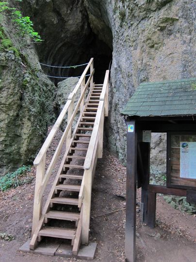 Holztreppe führt zum dunklen Eingang der Buchenlochhöhle. Rechts steht ein Infotafel mit Dach. Umgebung ist felsig und bewaldet.