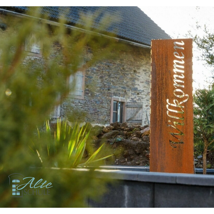 Ein rustikales Willkommensschild vor einem alten Gebäude. Umgeben von grünen Pflanzen und einem sonnigen Ambiente.